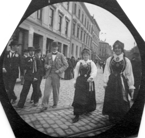Ludzie na ulicy, łącznie z kobietami w tradycyjnym, norweskim kostiumie. Jedna uśmiecha się niepewnie do kamery.